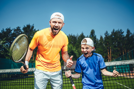 Portrait of shouting positive man and cheerful kid gesticulating hands while looking at camera. They holding rackets and standing on court for playing tennis outdoorの写真素材