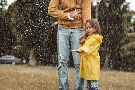 Full length side view of happy kid standing with father outside. She is stretching hand with content enjoying wet weather on nature with dadの写真素材