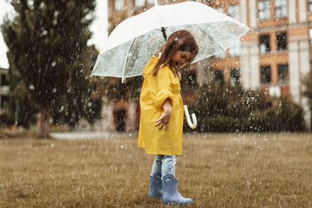 Full length of pleased kid playing on grass in rainy weather. She is standing with her back turned and glancing at rubber boots with smileの写真素材