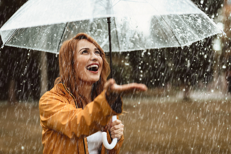 Waist up portrait of smiling girl standing outdoors with umbrella in hands. She is stretching hand and opening mouth in true pleasureの写真素材
