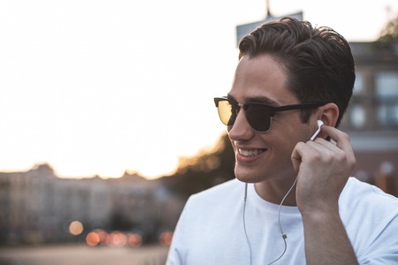 Side view of smiling young man standing on street with earphones. He is touching it with delight...