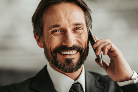 Portrait of beaming bearded businessman speaking on smartphone while looking at cameraの写真素材