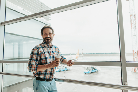 Portrait of beaming bearded man drinking cup of coffee while hearing song in airportの写真素材