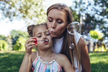 Waist up portrait of playful mother and small girl holding bottle with soap liquid. Delighted woman is blowing bubbles with joyの写真素材