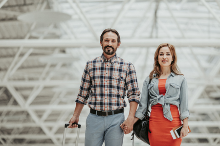 Portrait of cheerful bearded man and satisfied young girl holding hands together while going at airport before journey. She keeping documents and mobile in handの写真素材