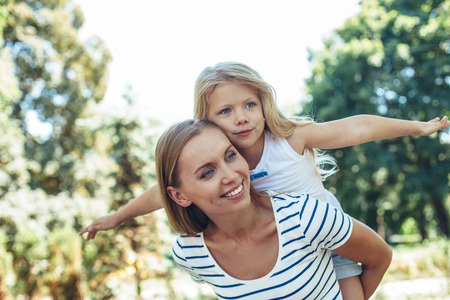 Waist up portrait of happy playful kid sitting on mom back with stretched hands to sides. She is pretending to fly while female is carrying her with content and smileの写真素材