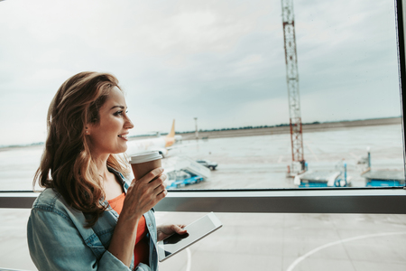 Side view satisfied girl tasting mug of beverage while holding electronic tablet in arm. She locating opposite indoor opposite airdromeの写真素材