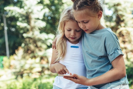 Waist up portrait of small girls standing in cuddles with phone in hands. They are playing games on smartphone peacefully. Copy space in left sideの写真素材