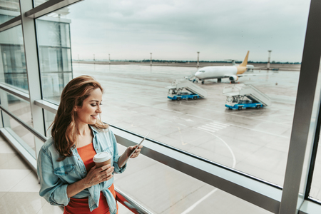 Side view happy lady watching at ticket while situating near window in airport. She drinking cup of delicious coffeeの写真素材