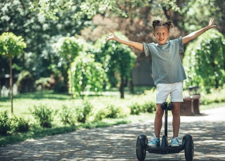 Full length portrait of content small kid riding self-balanced scooter. She is stretching hands to...