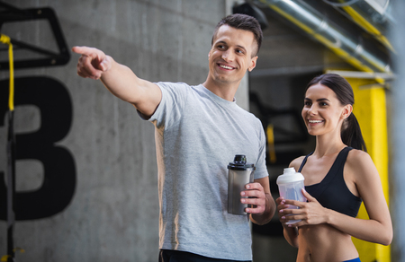 Smiling fit man is pointing at something while both he and his female friend are looking at it. Guy and young woman are standing and chatting during sport time in gym. Couple is using bottles with water for drinking during workoutの写真素材