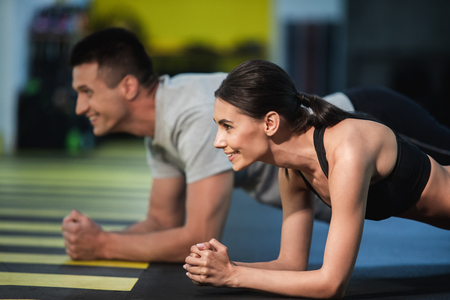 Focus on cheerful woman strained while doing plank. She is exercising with male friend and they are doing same static exercise while balancing on elbows. Two athletes are enjoying joint workout and feeling energeticの写真素材