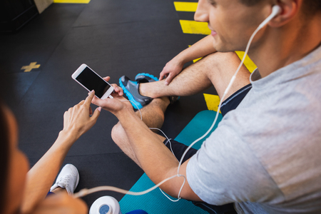 Top view of man holding smartphone while woman is pointing at screen. They are listening to music with earphones together. Friends are sitting on mats in sport studio during break in trainingの写真素材