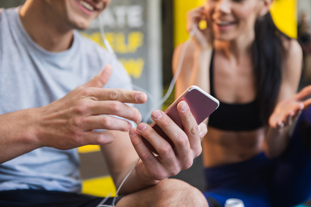 Focus on close-up male hands holding mobile phone. Cheerful man and woman are having fun while listening to same audio tracks with joint earphones in gym. They are sitting and chatting at same timeの写真素材