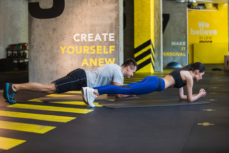 Athletic male and female are exercising together in gym. They are balancing on elbows while doing plank on mats. Couple training jointly indoors conceptの写真素材