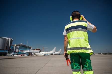 I hear you. Back view of man in signaling vest holding wands and checking the headset. Blue sky, runway, passenger planes and terminal on backgroundの写真素材