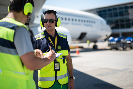 Right here. Smiling man in headphones pointing at clipboard while partner looking at writing data. Passenger plane on blurred backgroundの写真素材