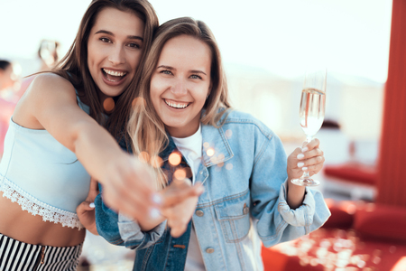 Portrait of smiling female hugging glad lady while keeping glass of champagne. Beaming friends having fun with bengal lights during partyの写真素材