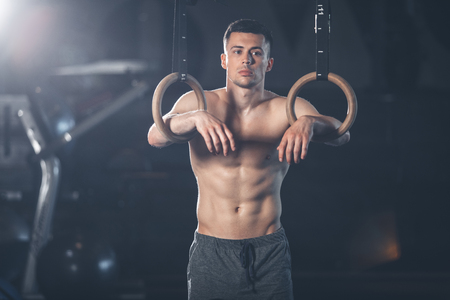 Brooding man is standing and leaning on sport rings. He is resting after acrobatic training with...