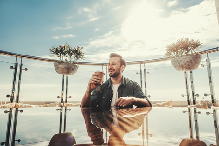 Lunch break. Bearded man holding coffee and smiling. He is sitting on the balcony of outdoor restaurantの写真素材