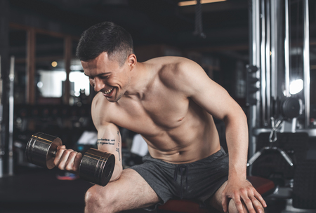 Cheerful guy is laughing while doing biceps curls. He is sitting on bench and exercising one arm by lifting dumbbell. Shirtless tattooed male is having fun during training in gymの写真素材