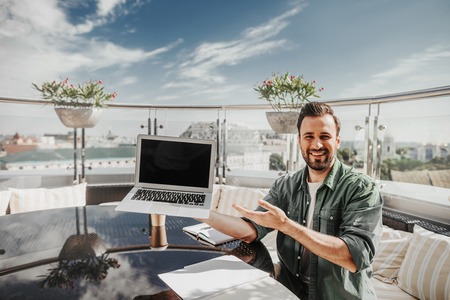 Look at this. Handsome cheerful guy representing notebook computer while sitting at outdoor cafeの写真素材