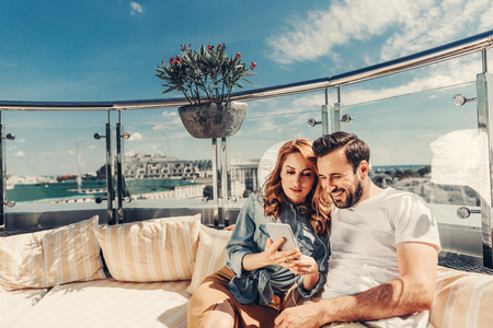 Excellent playlist. Smiling bearded man and lovely redhead lady sharing headphones and looking at mobile phone, They are sitting on the couch at outdoor cafeの写真素材