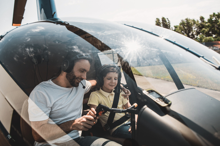 Beaming bearded dad teaching smiling kid to fly on rotorcraft. They locating inside of it while wearing headsets and communicatingの写真素材