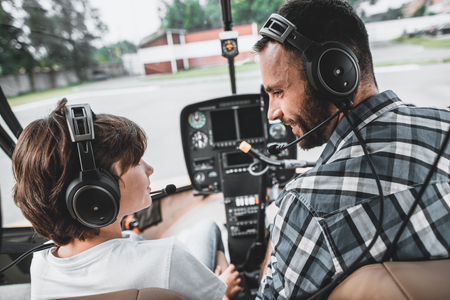 Side view smiling kid telling with cheerful bearded man while learning to control helicopter. They situating inside itの写真素材