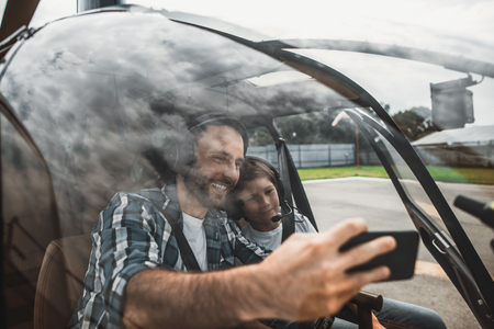 Portrait of happy bearded dad and outgoing kid taking selfie on mobile while wearing contemporary headsets. They having fun inside rotor plane with open door before flight. Glad parent and boy during shared hobby conceptの写真素材