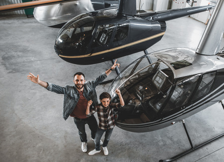 Full length top view portrait of cheerful bearded male and outgoing son flourishing arms while looking at camera. They standing near helicopters in garageの写真素材