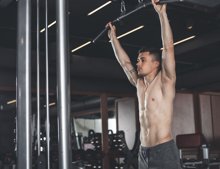 Shredded topless man is using pull down machine in gym. He is standing and holding handles while...