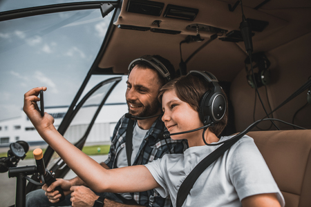 Side view smiling child and positive father taking photo on phone while wearing headphones. They locating in cabin of modern helicopterの写真素材