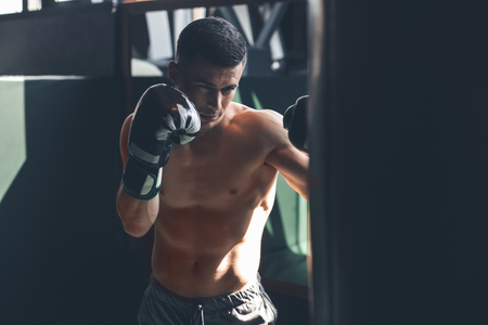 Shirtless shredded guy is having combat training in gym. He is kicking punching bag while wearing gloves. Male is concentrated on fightingの写真素材