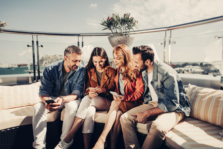 Nice meeting. Two smiling bearded men and beautiful young ladies using mobile phone at rooftop cafeの写真素材