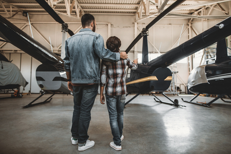 Full length kid pointing hand on big rotor plane. Man hugging him while listening his description of vehicle. They turning back to cameraの写真素材