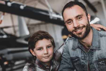 Portrait of beaming kid embracing cheerful unshaven dad while they looking at camera. They locating indoor. Positive father having leisure with son conceptの写真素材