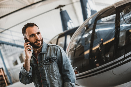 Portrait of outgoing unshaven male speaking on mobile while locating near helicopters inside garage