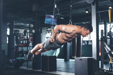 Shredded guy is feeling tension while exercising with gymnastic equipment. He is straightening all body and bringing it to parallel to floor. Male is holding handles and looking downの写真素材
