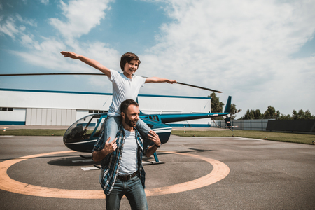Portrait of smiling son playing with optimistic dad while sitting on his shoulders and gesticulating hands. He imaging fly. They locating near helicopter on landing fieldの写真素材