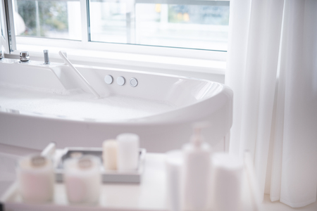 Close up white beautiful bath with clean water locating in bright cozy apartment for treatment and spa procedures. Different bottles with liquid situating opposite itの写真素材