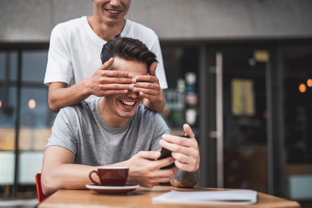 Who is it. Cheerful guy closing face of laughing comrade. He sitting at table and typing in modern mobileの写真素材