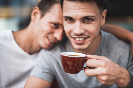 Beaming guy tasting appetizing mug of liquid while talking with happy friend. He leaning on him. They looking at cameraの写真素材