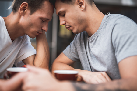 Side view calm male touching forehead of another serene guy while tasting cups of appetizing liquid at deskの写真素材