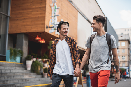 Cheerful male keeping arm of positive guy while walking outdoor. They looking at each other during conversationの写真素材