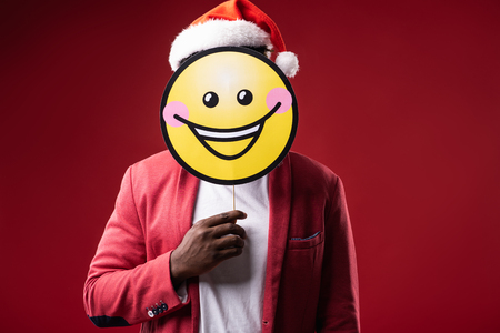 Happy emotion. Man in santa hat hiding under carton icon of smiling face. Isolated on red backgroundの写真素材