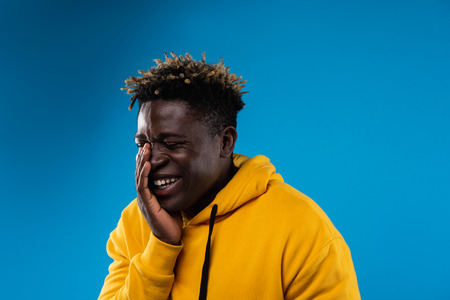 Funny moment. Portrait of young male with closed eyes chuckling at something. He is wearing yellow hoodie and standing on blue backgroundの写真素材