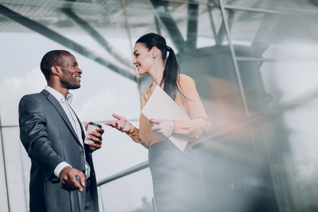 How are things. Waist up portrait of business lady holding folder and talking with her colleague, while drinking coffee. They are smiling looking at each otherの写真素材