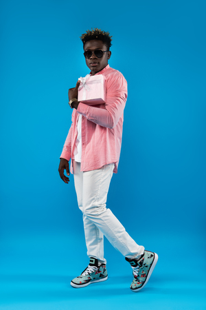 Best wishes. Full length portrait of young man in sunglasses holding pink gift box with ribbon. Isolated on blue backgroundの写真素材