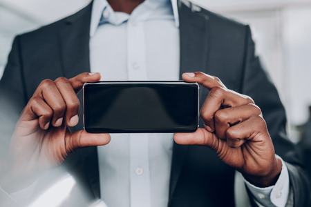 My new gadget. Close up of male hands taking cellphone. Smiling guy is standing in the office room and showing gadgetの写真素材
